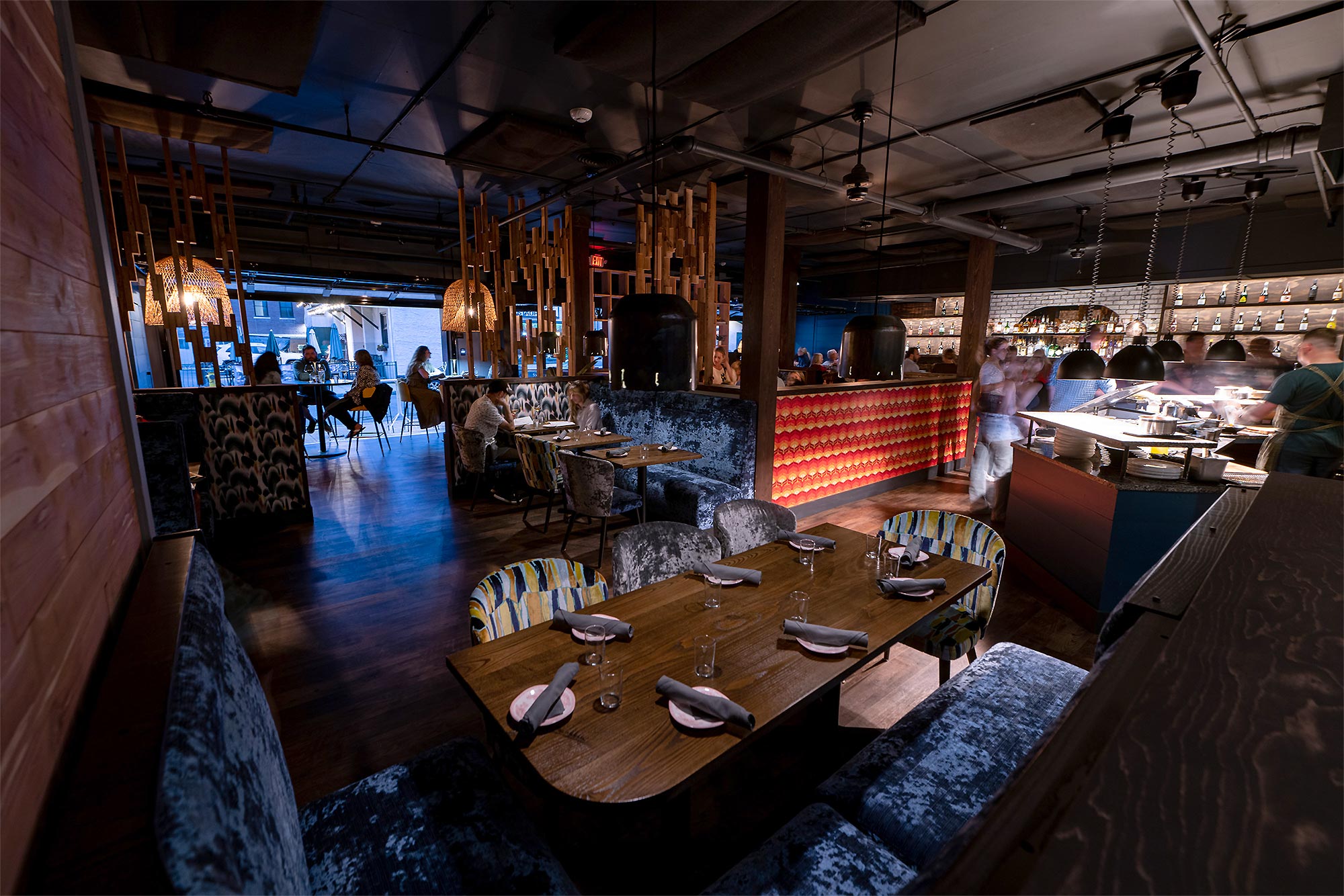Wide angle view of restaurant seating with patrons at tables, chefs in the open kitchen, and the bar in the background